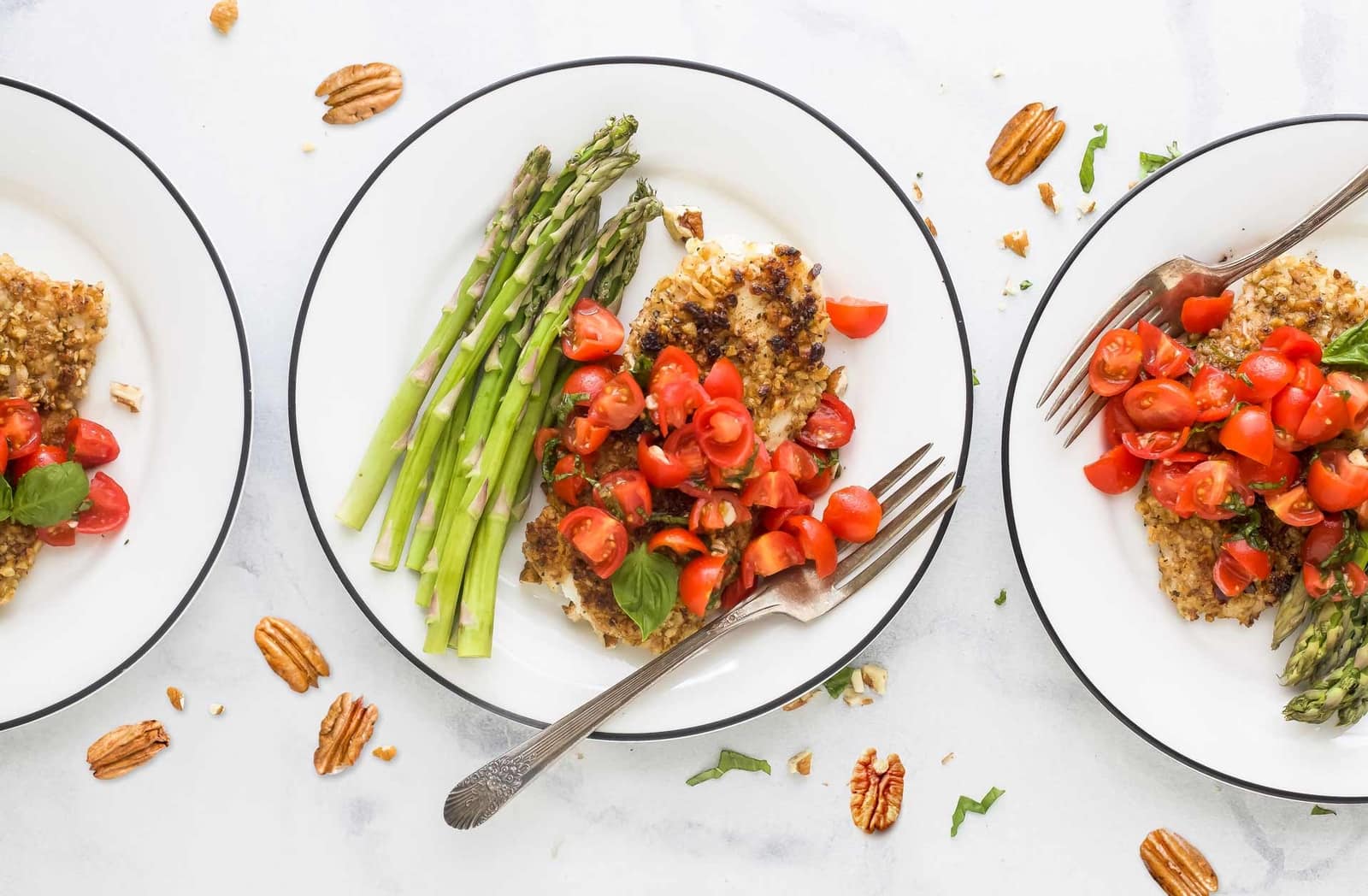 Plates of pecan-crusted tilapia topped with fresh tomato bruschetta and served with tender asparagus spears, styled on a light surface with scattered pecans and basil for garnish