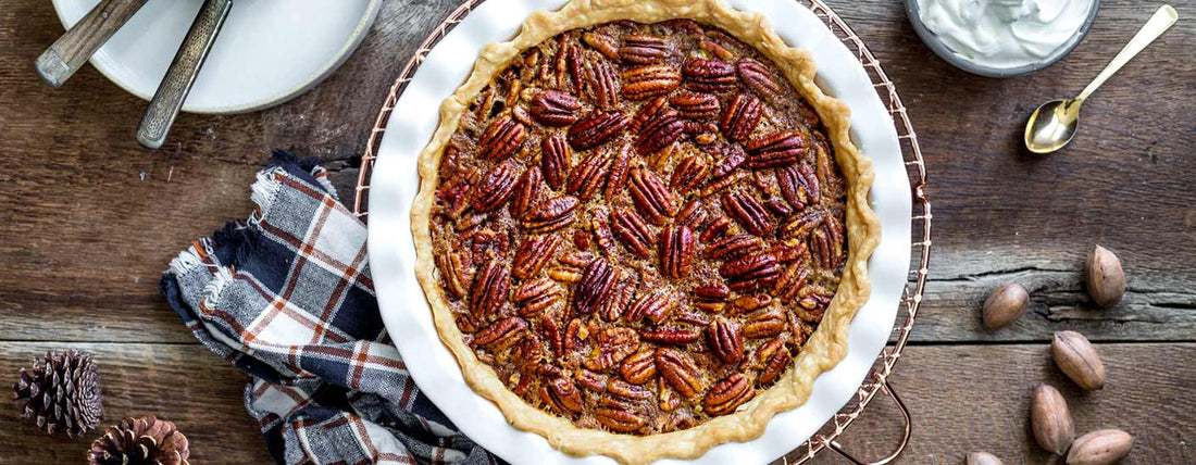 A freshly baked classic pecan pie in a white ceramic dish, its glossy, golden-brown pecans arranged neatly on top, set on a rustic wooden table with a plaid napkin, pinecones, and serving utensils nearby