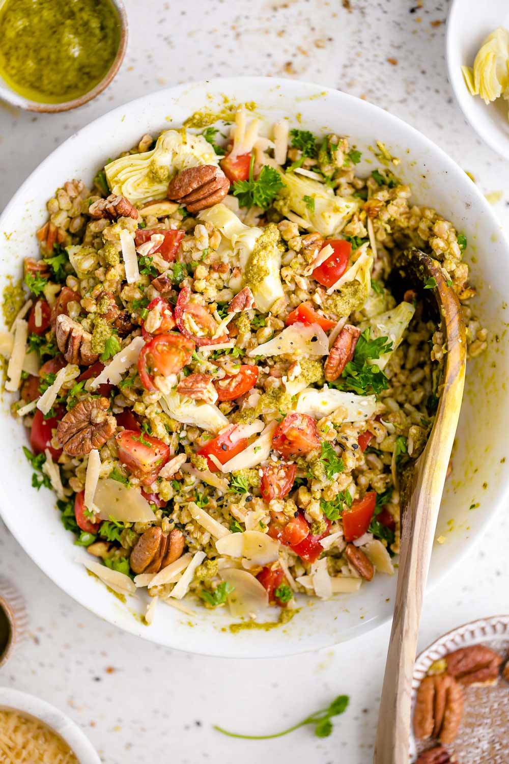 A vibrant farro salad tossed with cherry tomatoes, pecans, artichoke hearts, shaved Parmesan, and fresh herbs, drizzled with green pesto and served in a white bowl with a wooden spoon