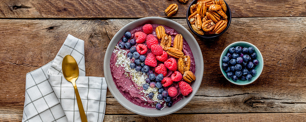 A vibrant acai smoothie bowl topped with fresh raspberries, blueberries, shredded coconut, and crunchy pecan halves, styled on a rustic wooden table beside a gold spoon and small bowls of extra toppings