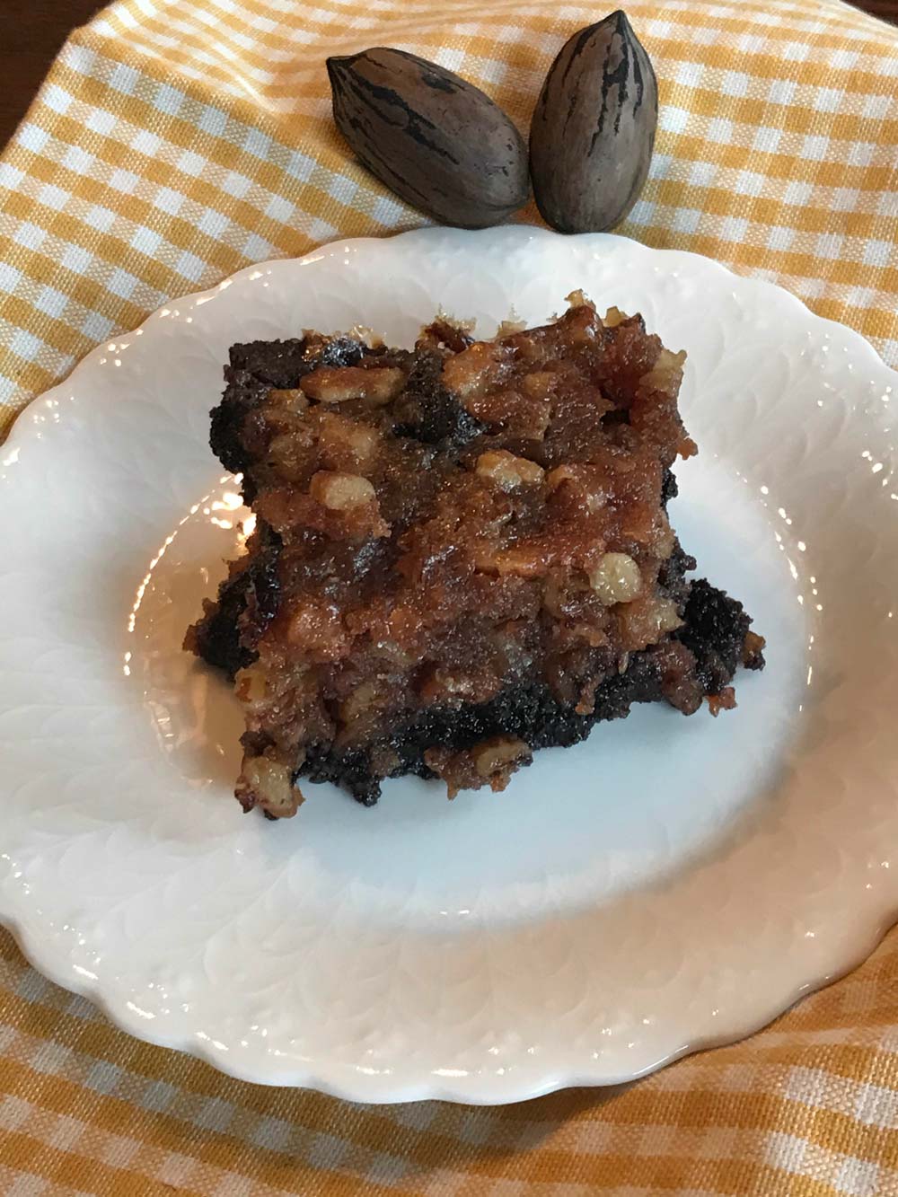 A rich pecan brownie served on a white plate over a yellow checkered cloth, topped with a gooey pecan and caramel layer, with two whole in-shell pecans placed in the background for garnish
