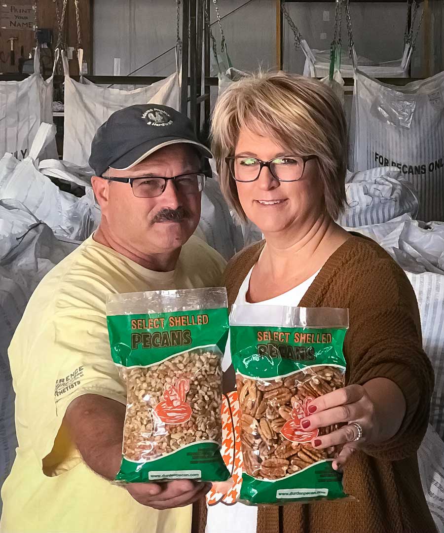 A man and woman standing inside a pecan processing facility, proudly holding up two bags of Durden Pecan Co. select shelled pecans—halves and pieces—ready for customers